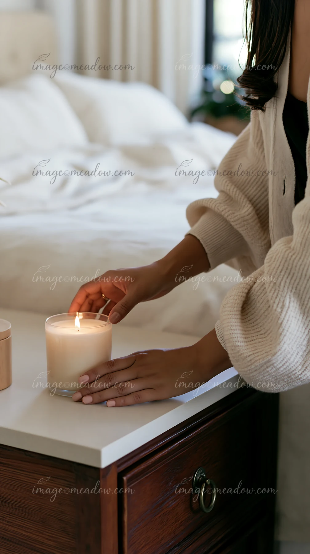 Hands light a vanilla candle placed on a bedside table in a softly lit room.