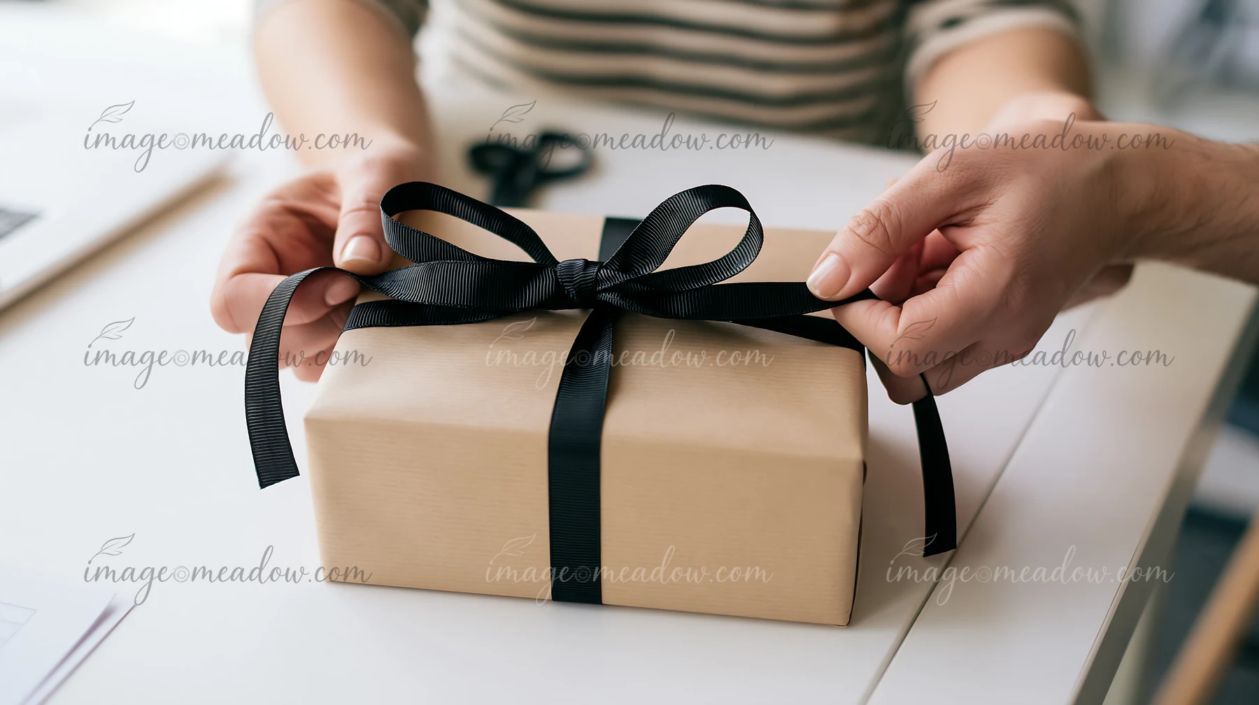 Hands tie a black ribbon around a kraft paper gift box on a white tabletop.