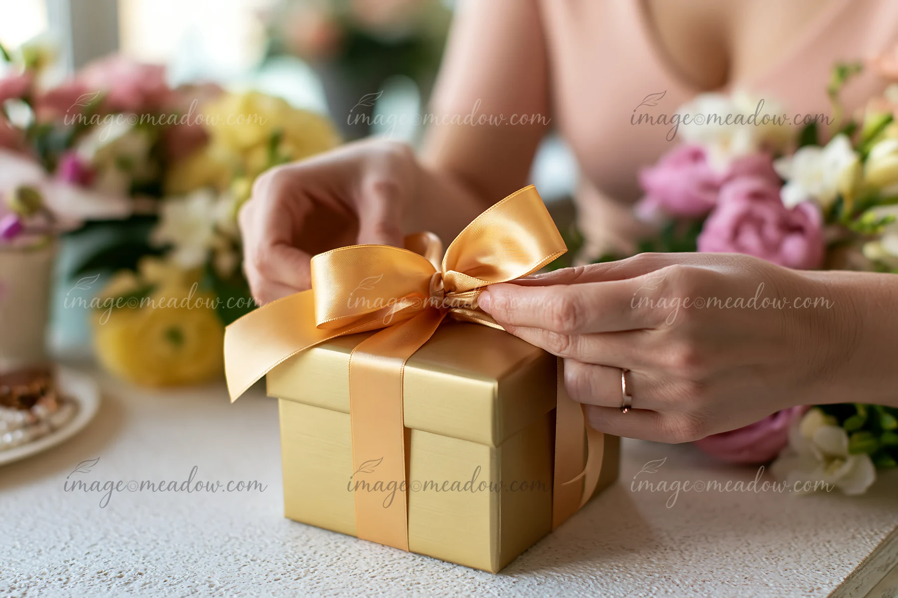 Hands tie a gold ribbon around a gift box on a table, surrounded by colorful flowers.