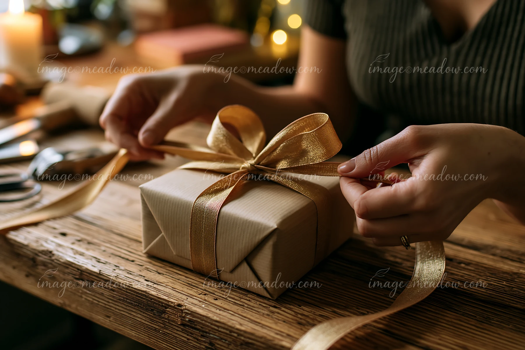 Hands tie a gold ribbon around a wrapped gift box on a rustic wooden table.