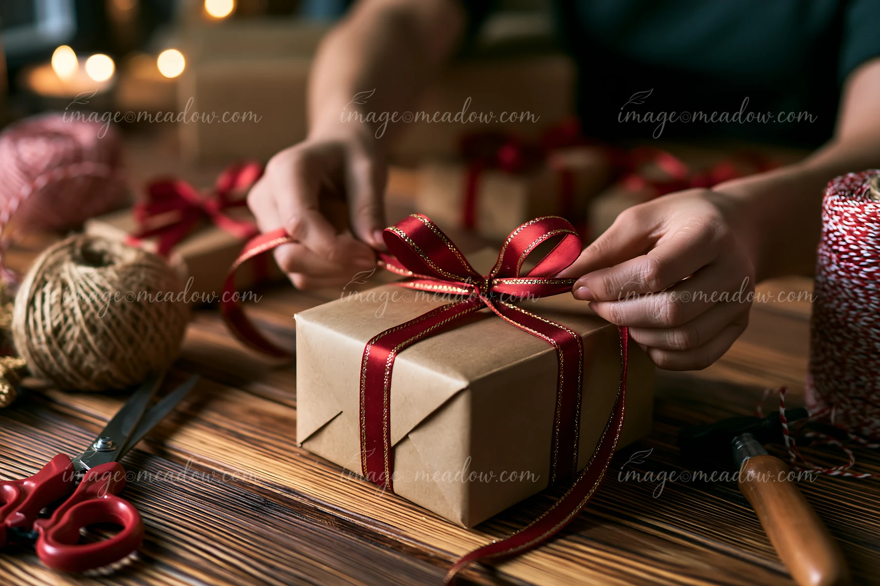Hands tie a red ribbon around a wrapped gift box on a wooden worktable.