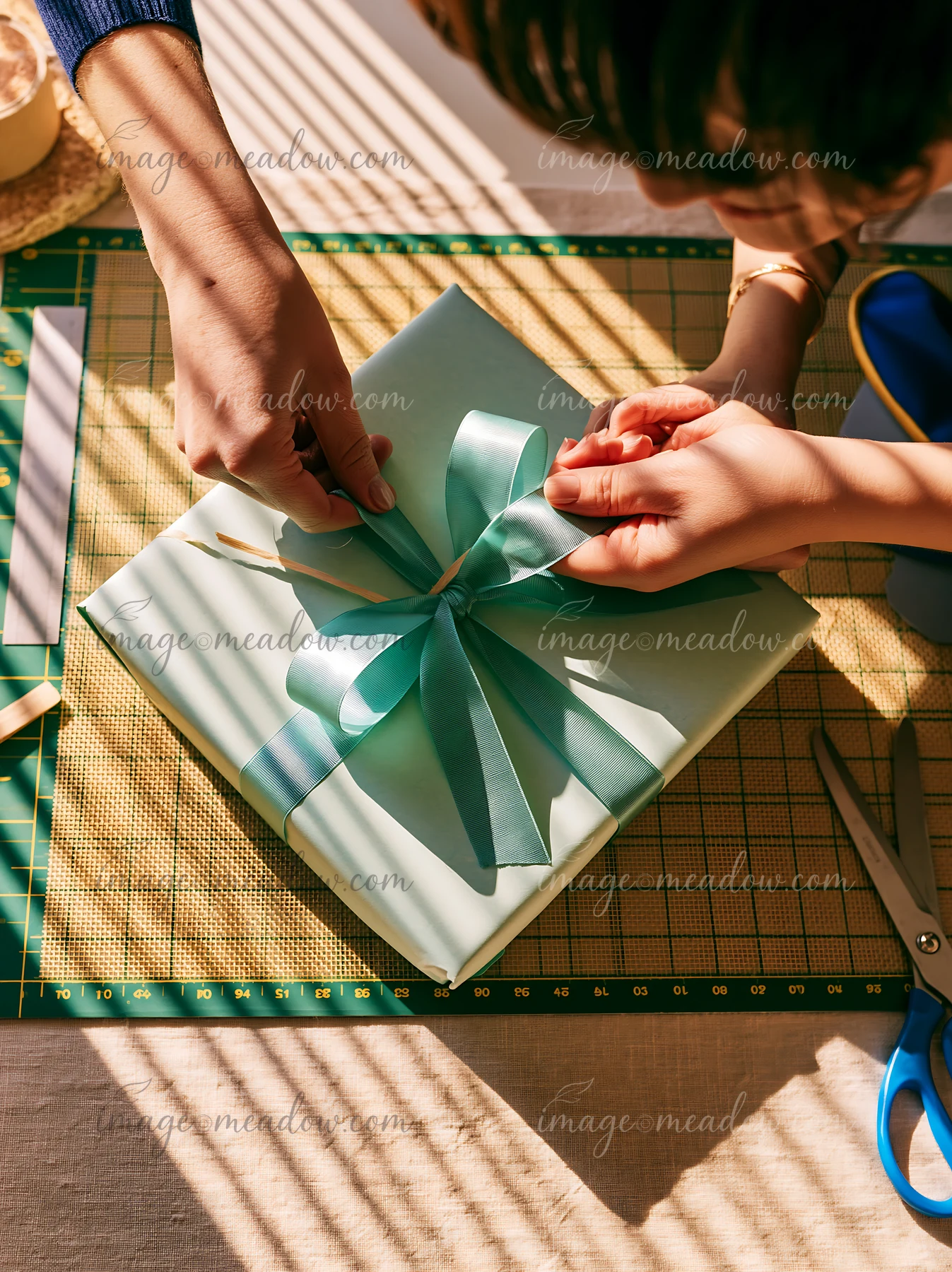 Hands tie a teal ribbon around a wrapped gift box on a sunlit crafting table.