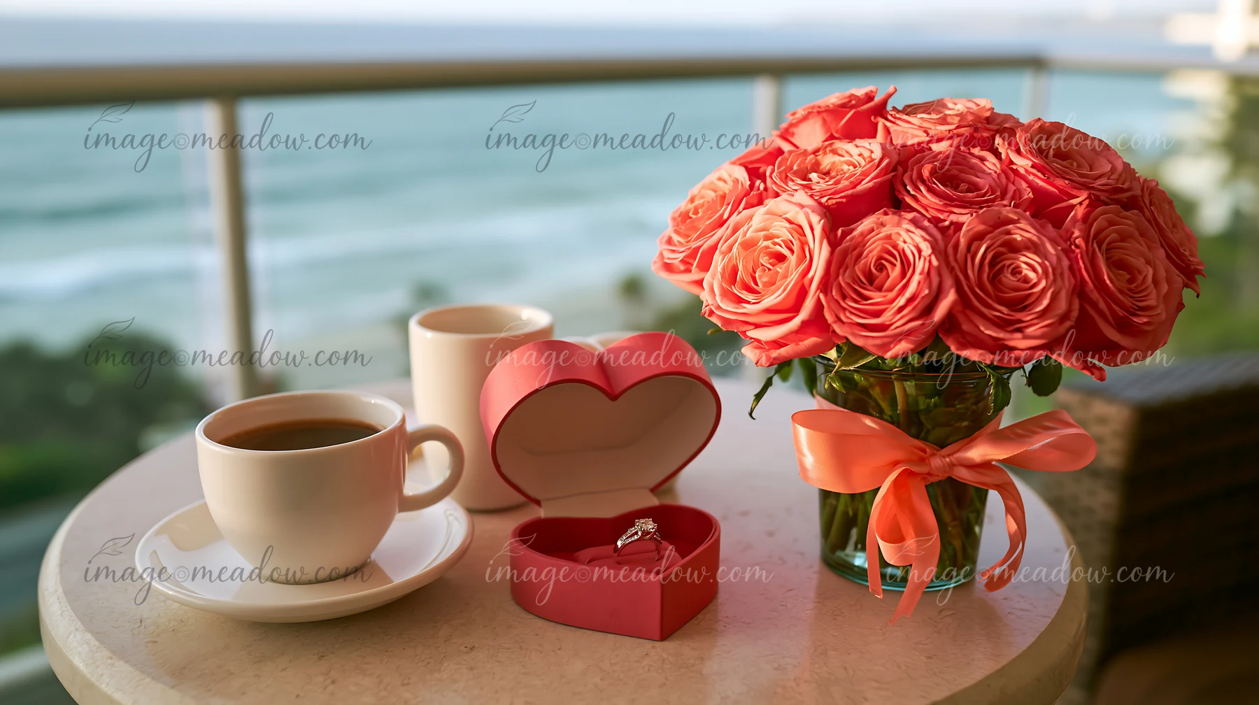 Heart-shaped ring box beside coral roses on a table with coffee cups and a blurred ocean view.