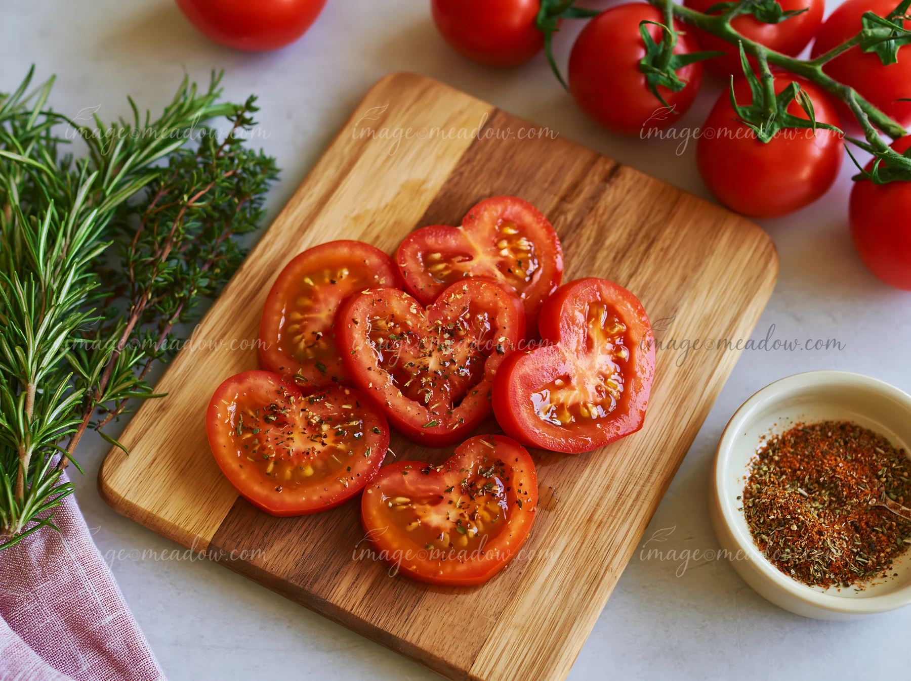 Heart-shaped tomato slices are arranged on a wooden cutting board, surrounded by fresh herbs and whole tomatoes.