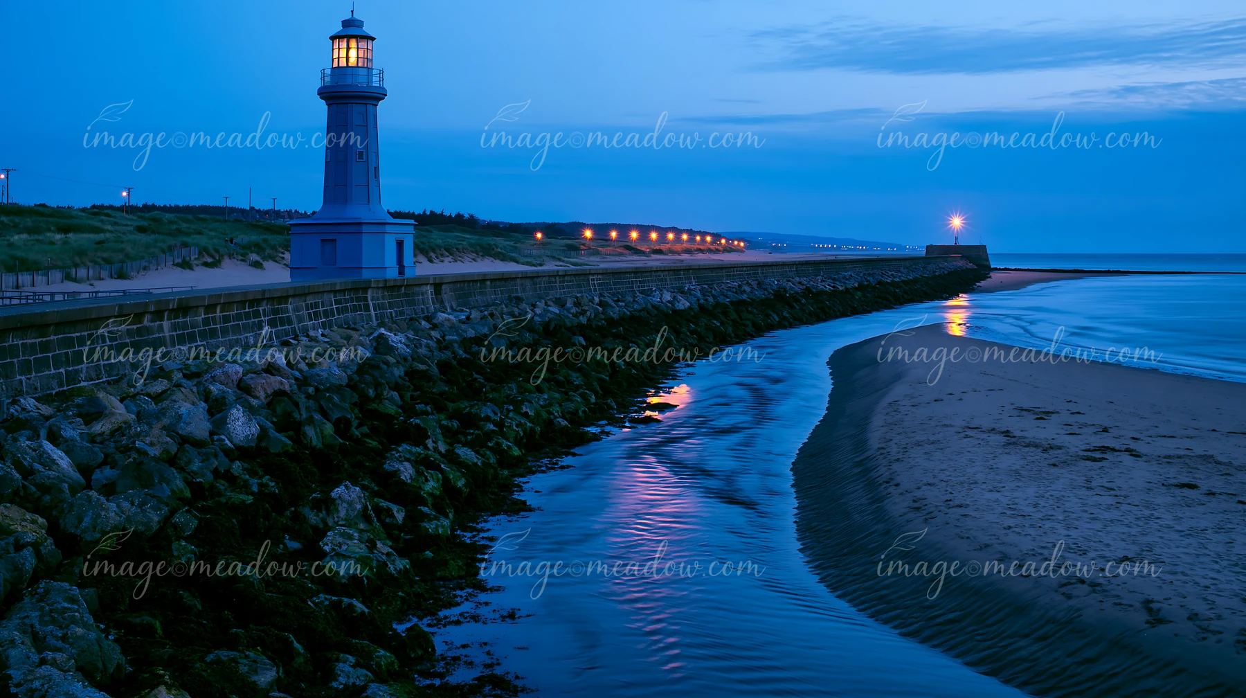 Illuminated lighthouse beside a rocky seawall and calm shoreline at blue hour.