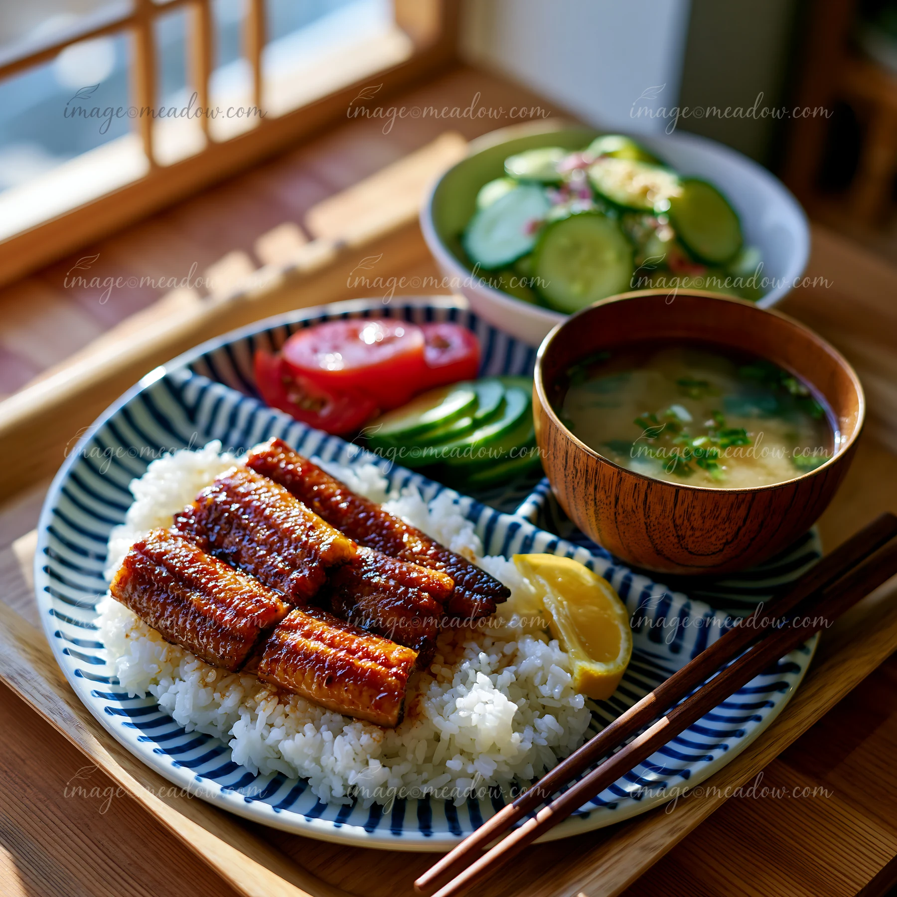 Japanese-style meal with glazed grilled eel over rice, soup, salad, lemon, and chopsticks on a sunlit wooden table.