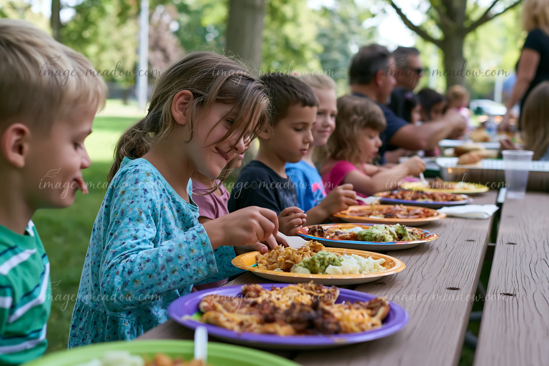 Kids eat from colorful plates at a charity event buffet held outdoors in a park.
