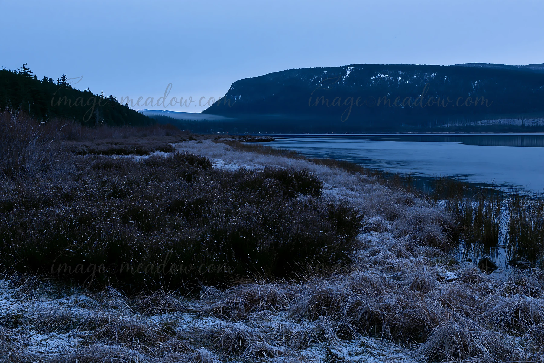Snow covers a meadow beside a lake. The distant hills show signs of winter. The sky is darkening as evening approaches. The scene captures the quietness of natu