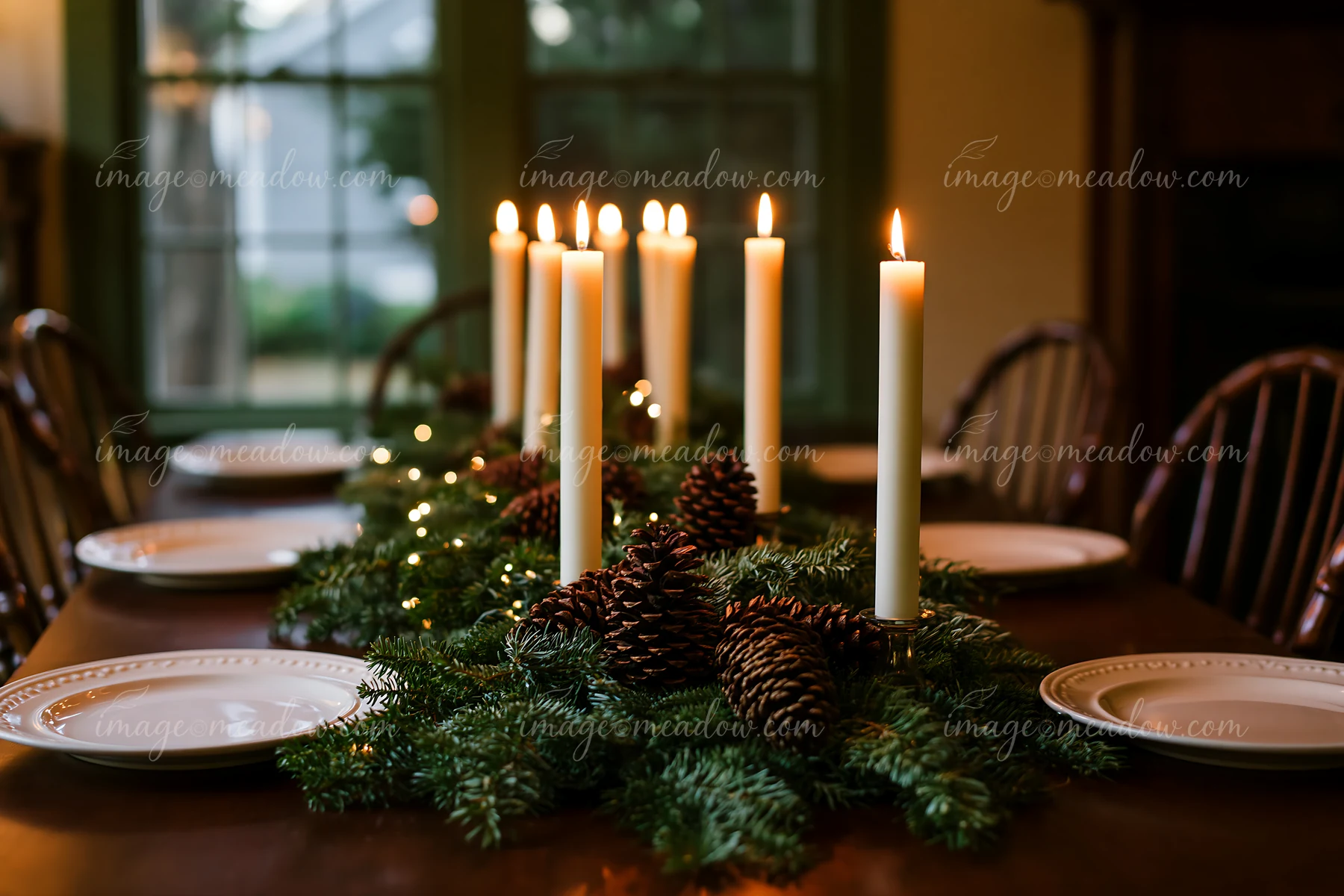 Lit candles, evergreen branches, and pine cones form a Christmas centerpiece on a dining table with open space around it.