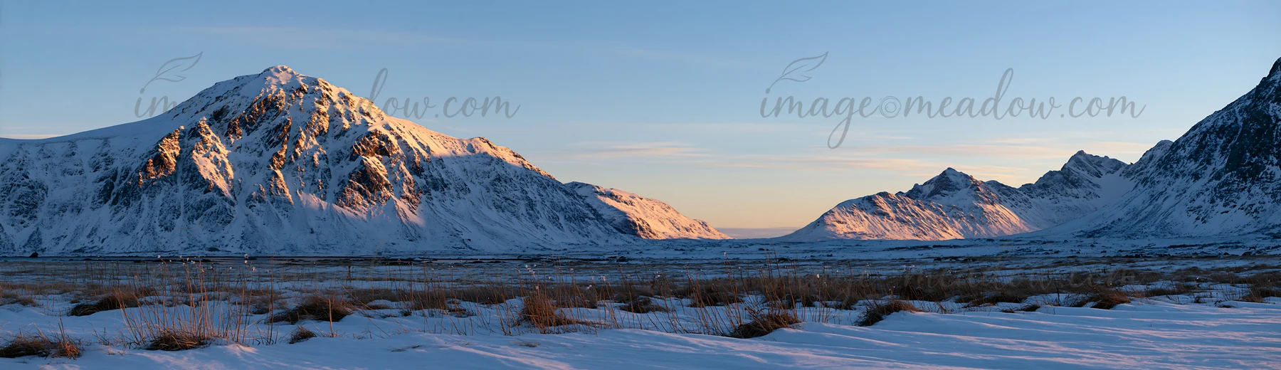 The scene shows snow-covered meadows in Lofoten, surrounded by mountains under a clear blue sky during winter. The landscape highlights the rugged terrain and d