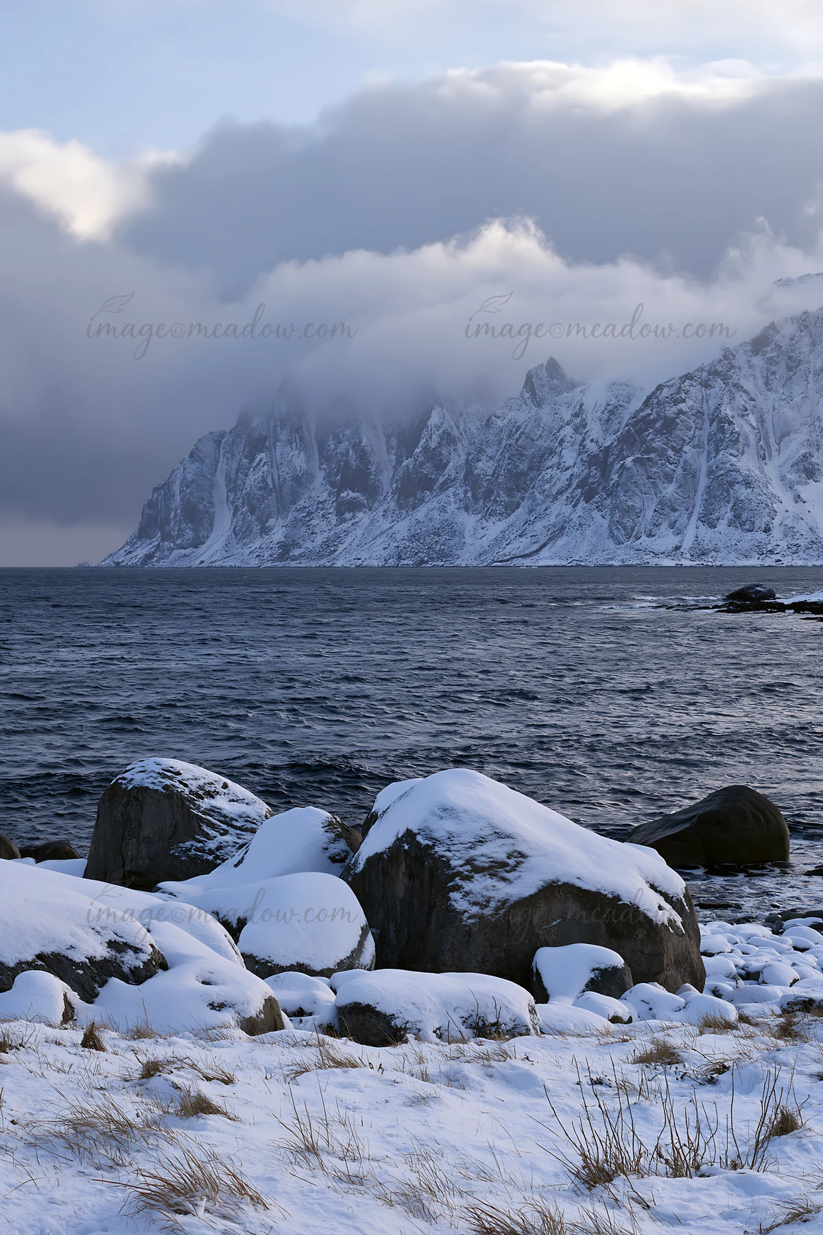 A snowy coastal meadow in Lofoten, with large rocks at the shore and mountains in the background, under cloudy skies on a winter day.