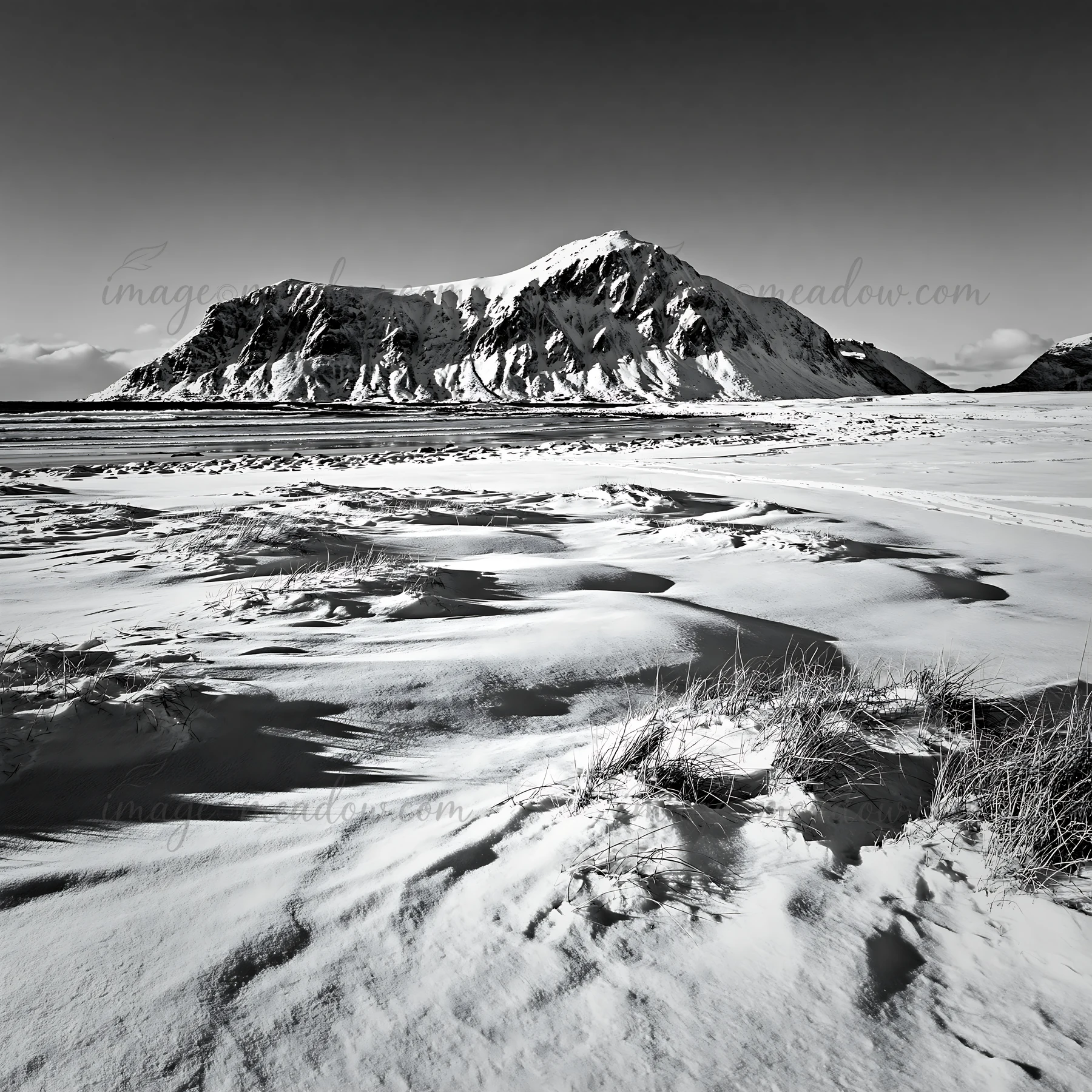 Snow blankets the coastal meadow in Lofoten, with mountains in the background. The scene captures the stark contrasts of black and white. Winter conditions crea