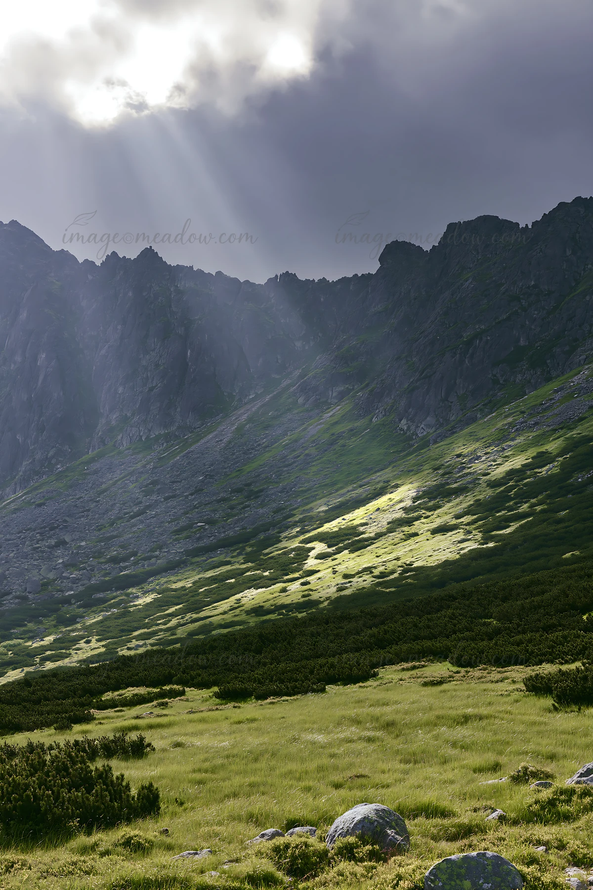 Sunrays break through clouds, illuminating a green meadow against a rocky mountain. The scene captures nature's beauty at a high elevation with patches of sunli