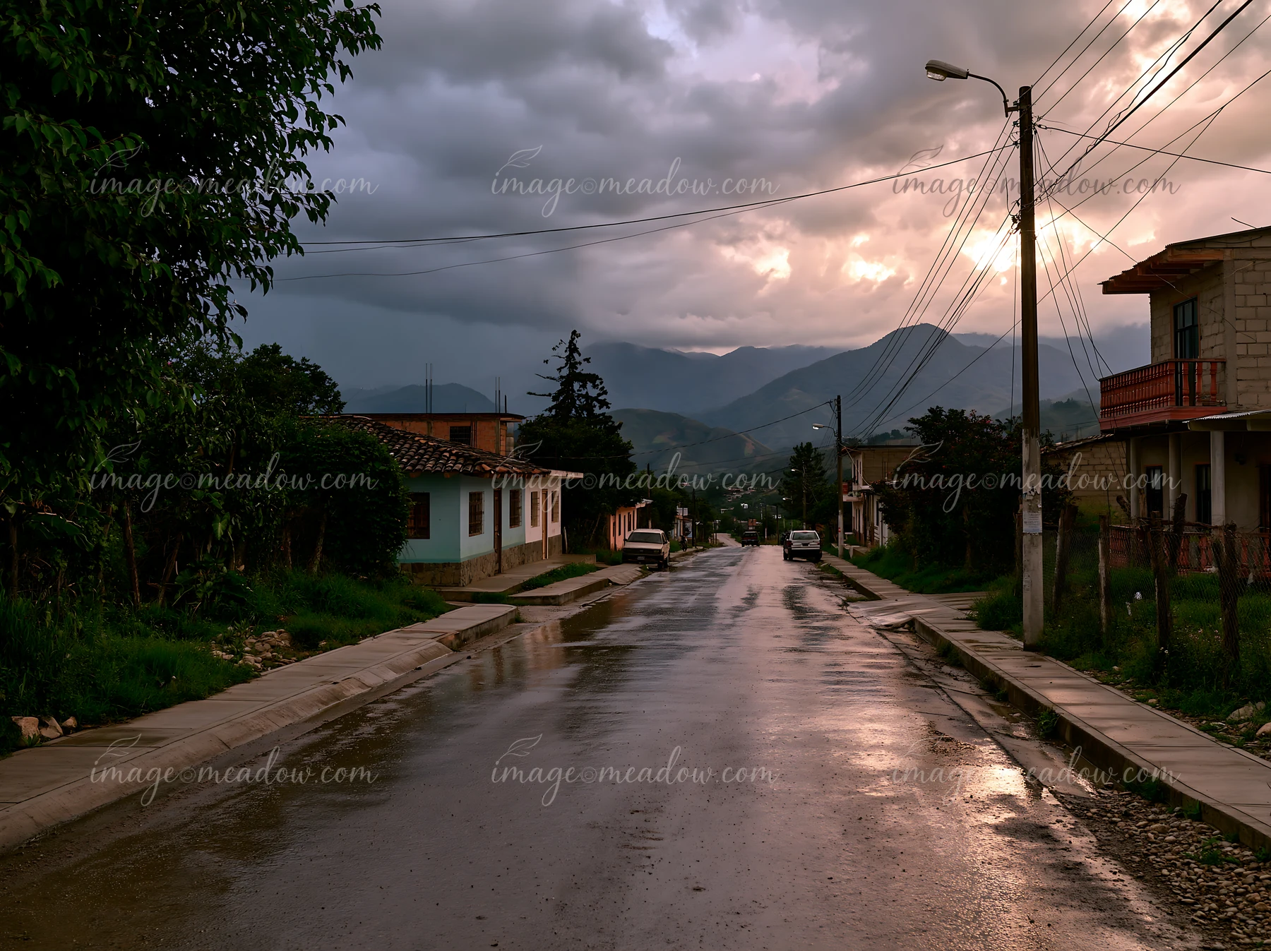 Moody clouds gather over a mountain range as rain falls, creating reflections on the wet street.