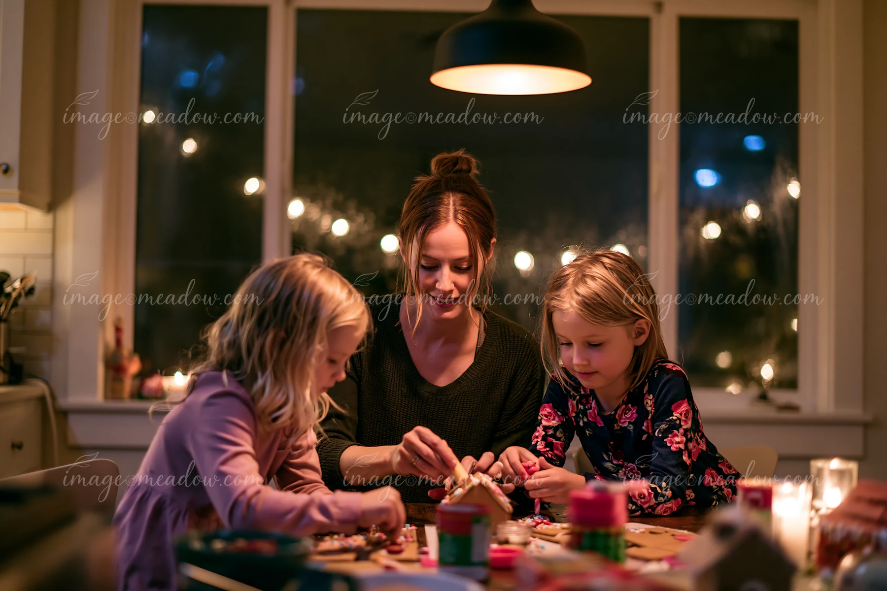 Mother and two daughters assemble and decorate a gingerbread house at a warmly lit dining table.