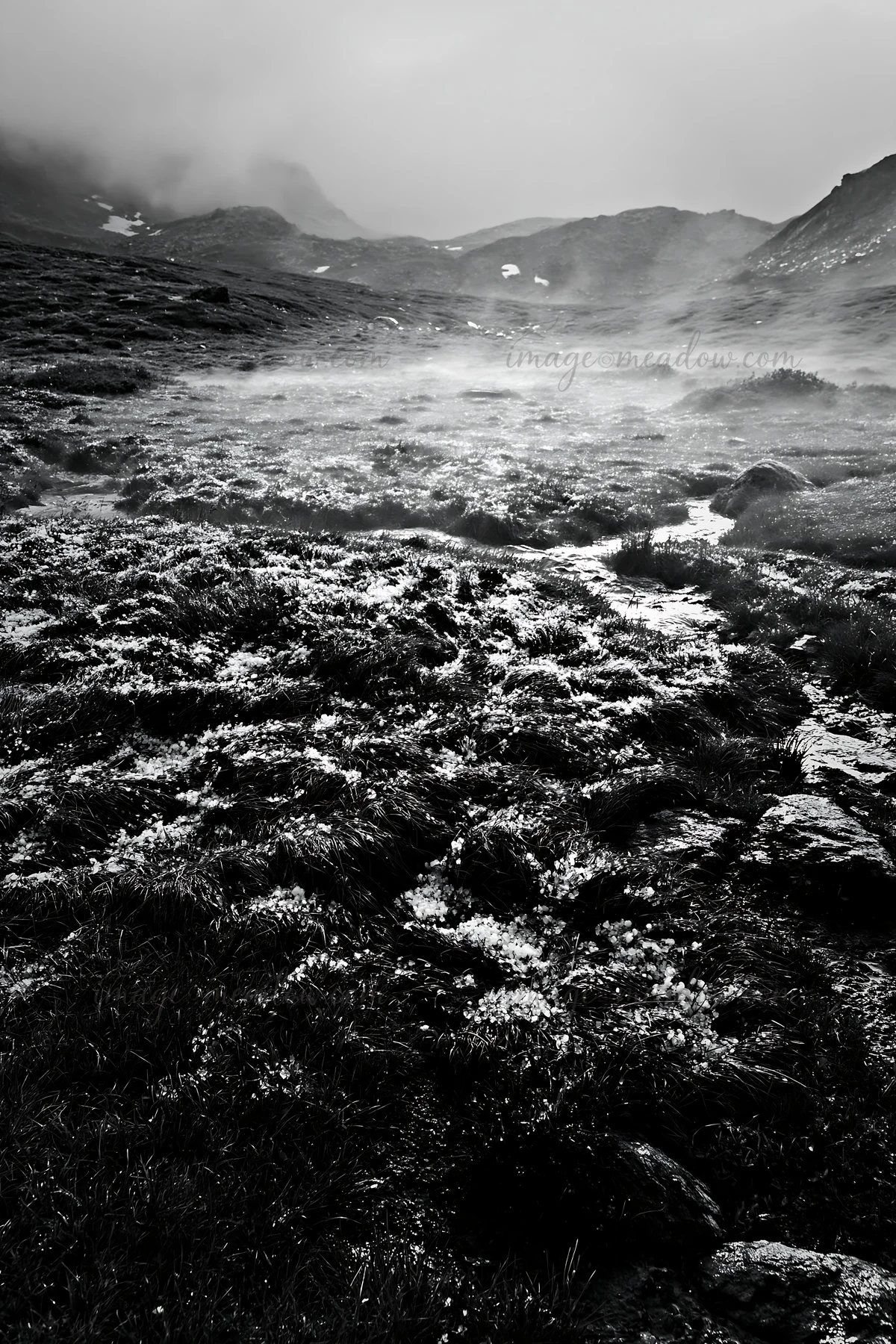 A view of a mountain pass meadow shows thick grass with patches of snow and mist rising from the ground. The landscape features rocky areas and distant peaks un