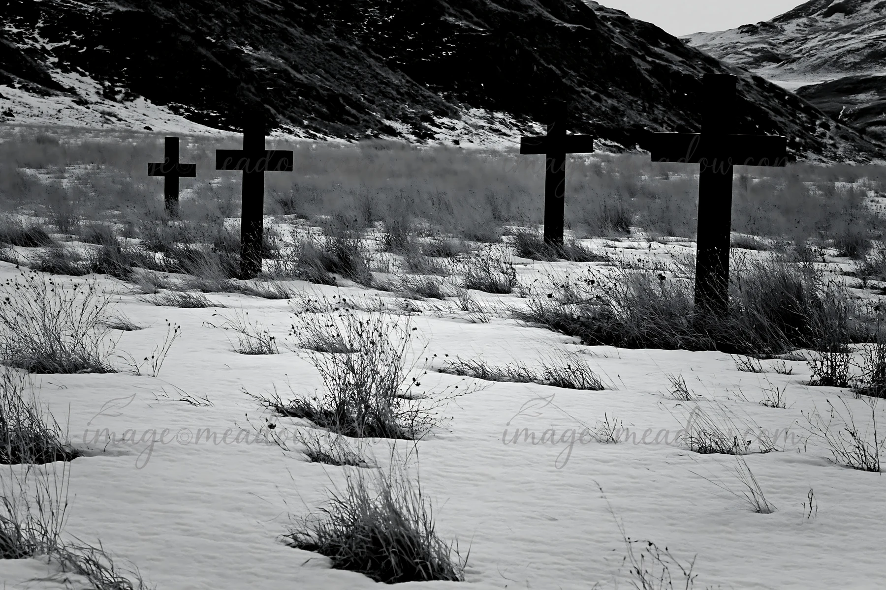 Multiple Christian crosses stand in a snowy meadow with sparse grass. The scene has a grey sky and distant hills, highlighting the crosses against the winter la