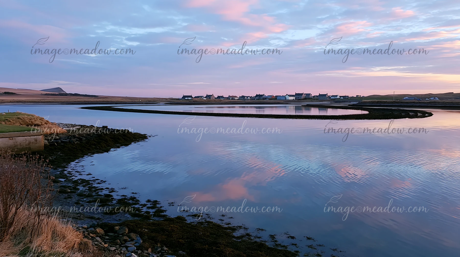 Mussel farm structures extend across calm coastal water beside a village beneath a pastel sunset sky.