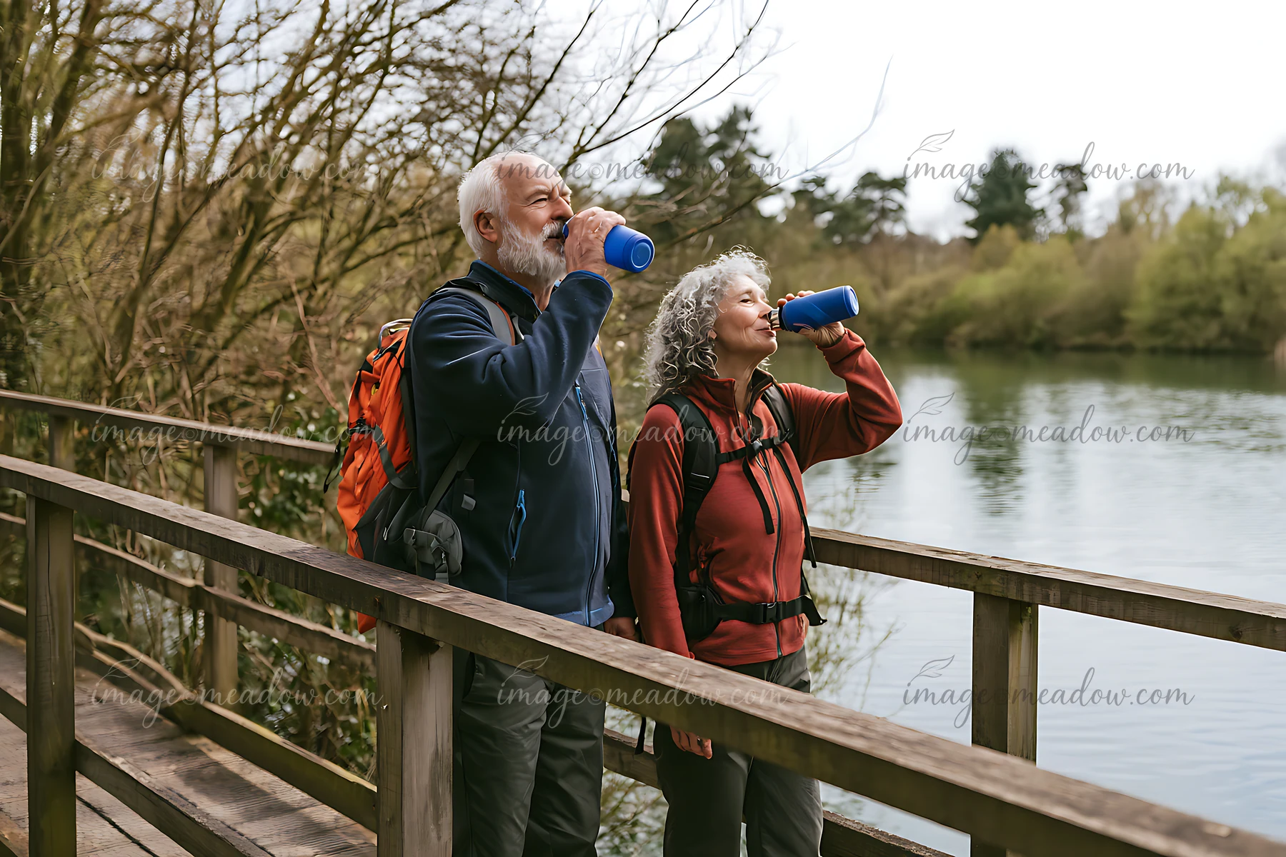 Older couple drinks water while hiking across a wooden footbridge beside a calm lake.