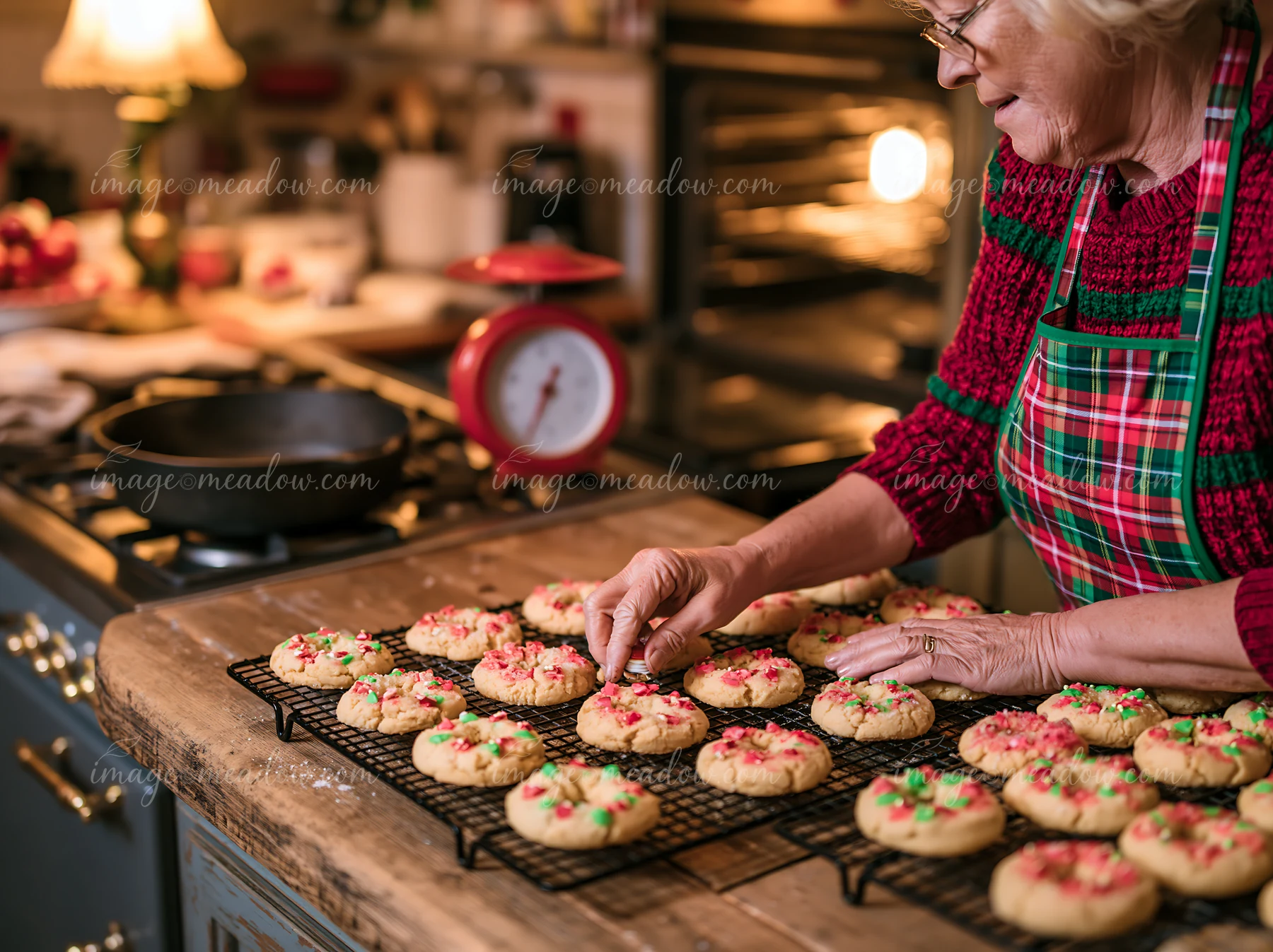 Older woman arranges colorful Christmas cookies cooling on wire racks in a warmly lit kitchen.