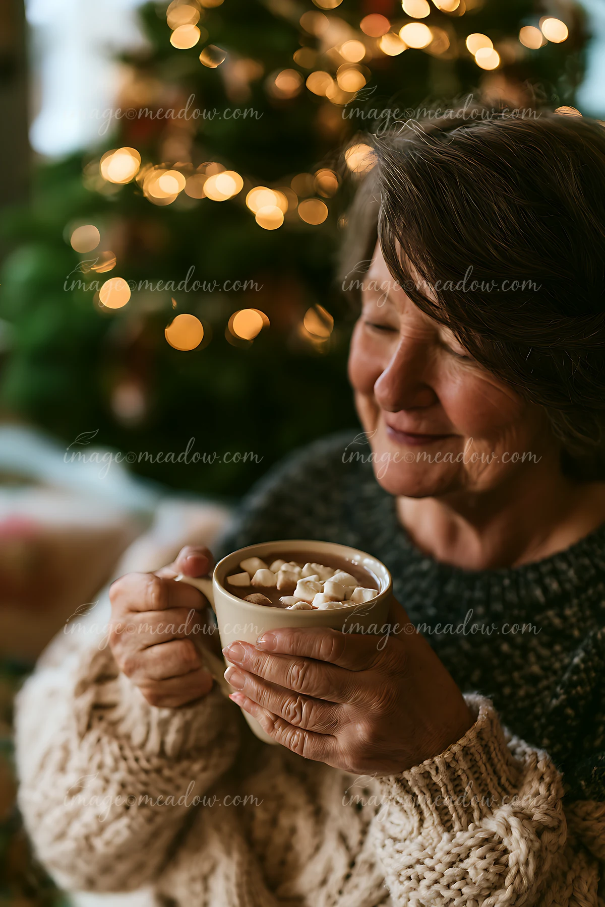 Older woman holding a mug of cocoa with marshmallows beside a softly lit evergreen tree.