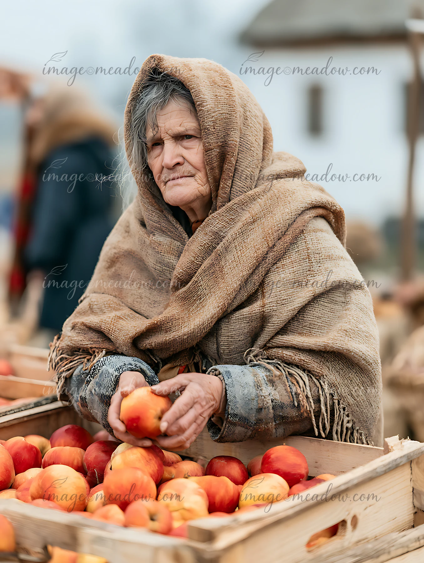 Older woman in a woven shawl holds an apple beside wooden crates filled with apples at an outdoor market.