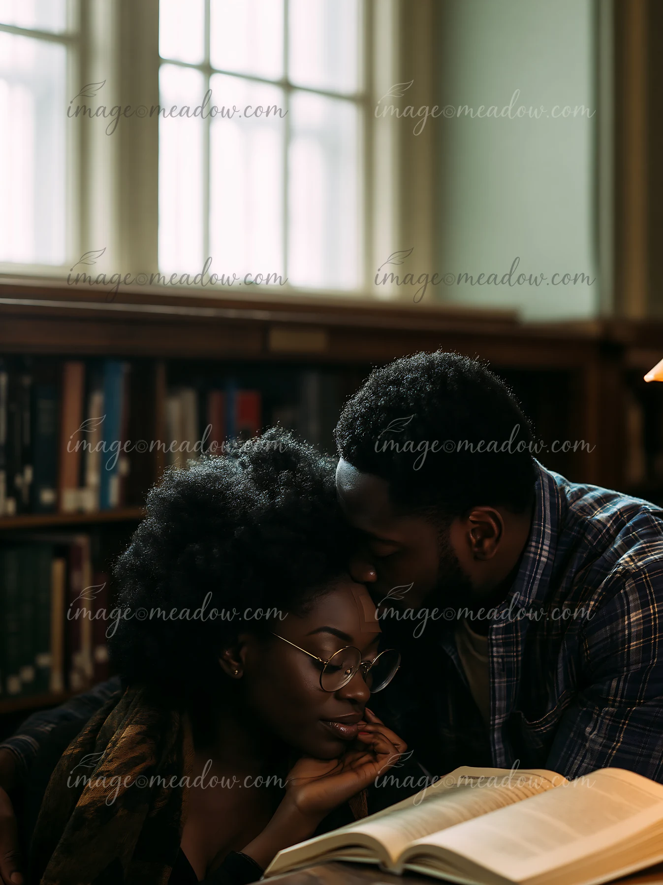 One partner kisses the other’s forehead beside an open book in a warmly lit library.