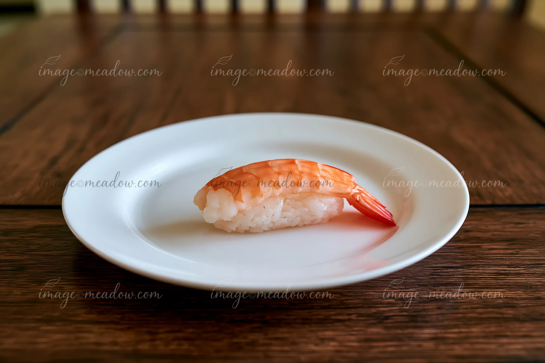 One shrimp sushi piece on a white plate sits on a wooden table.