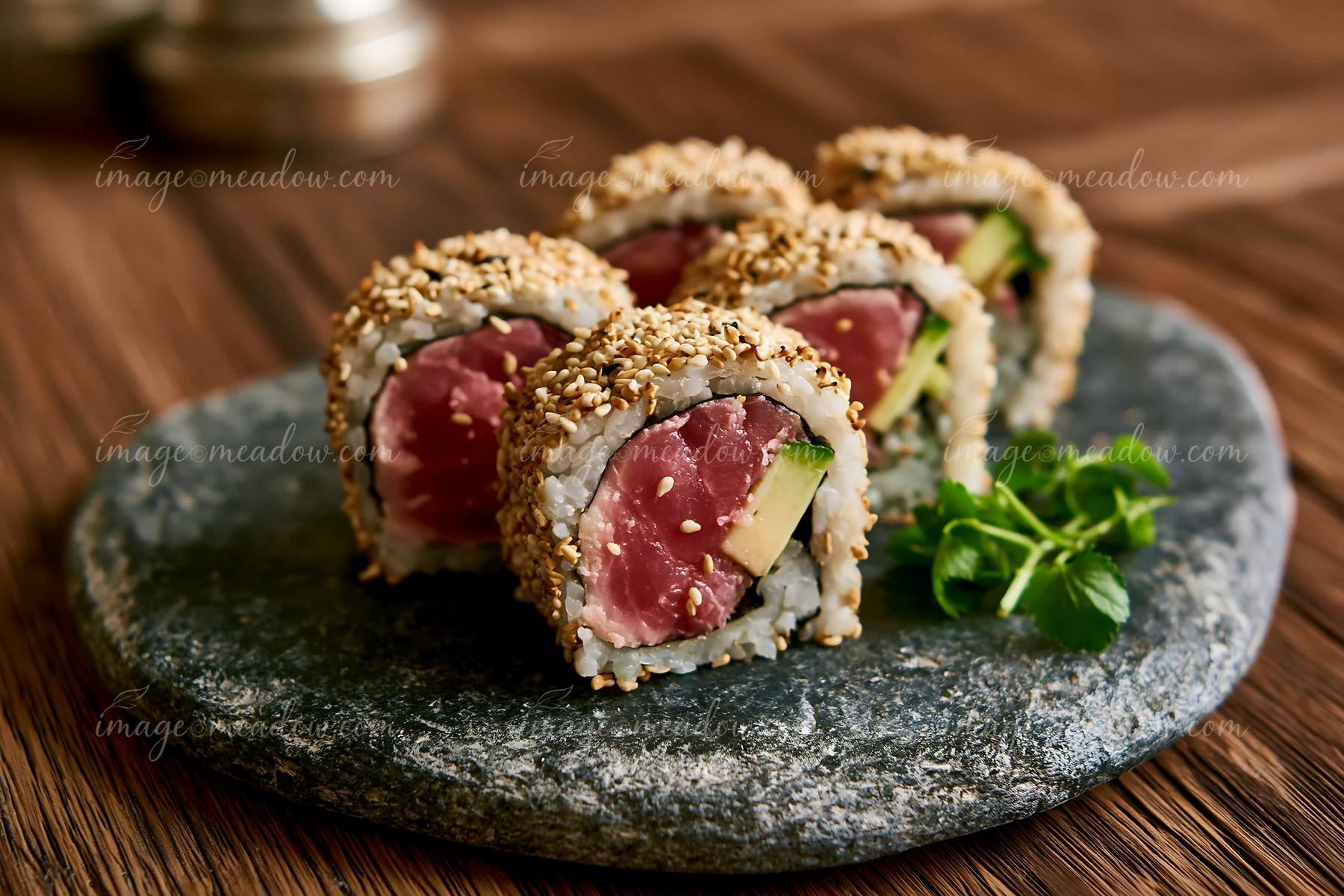 One tuna roll slice with sesame coating is placed on a dark stone plate alongside three other sushi pieces.