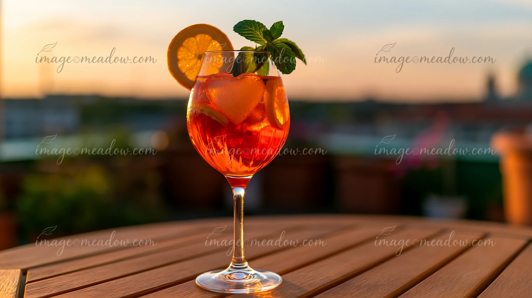 Orange cocktail with a heart-shaped ice cube, citrus garnish, and mint on a wooden table at sunset.
