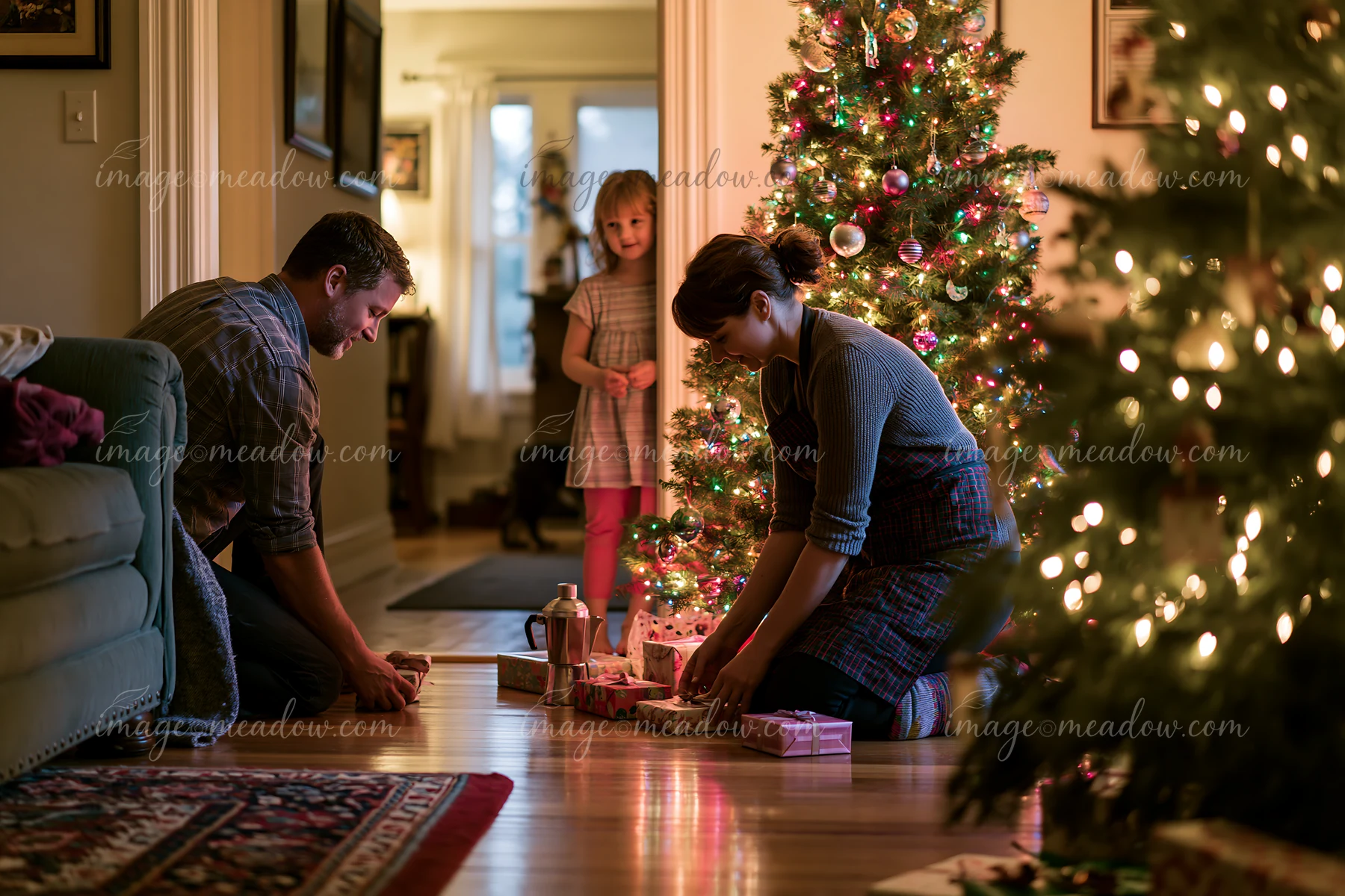 Parents are placing gifts under a Christmas tree in a cozy living room while their child looks on with curiosity.