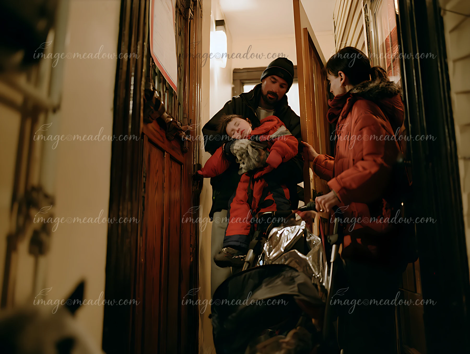 Parents carry a sleeping child in a stroller through a warmly lit interior hallway.