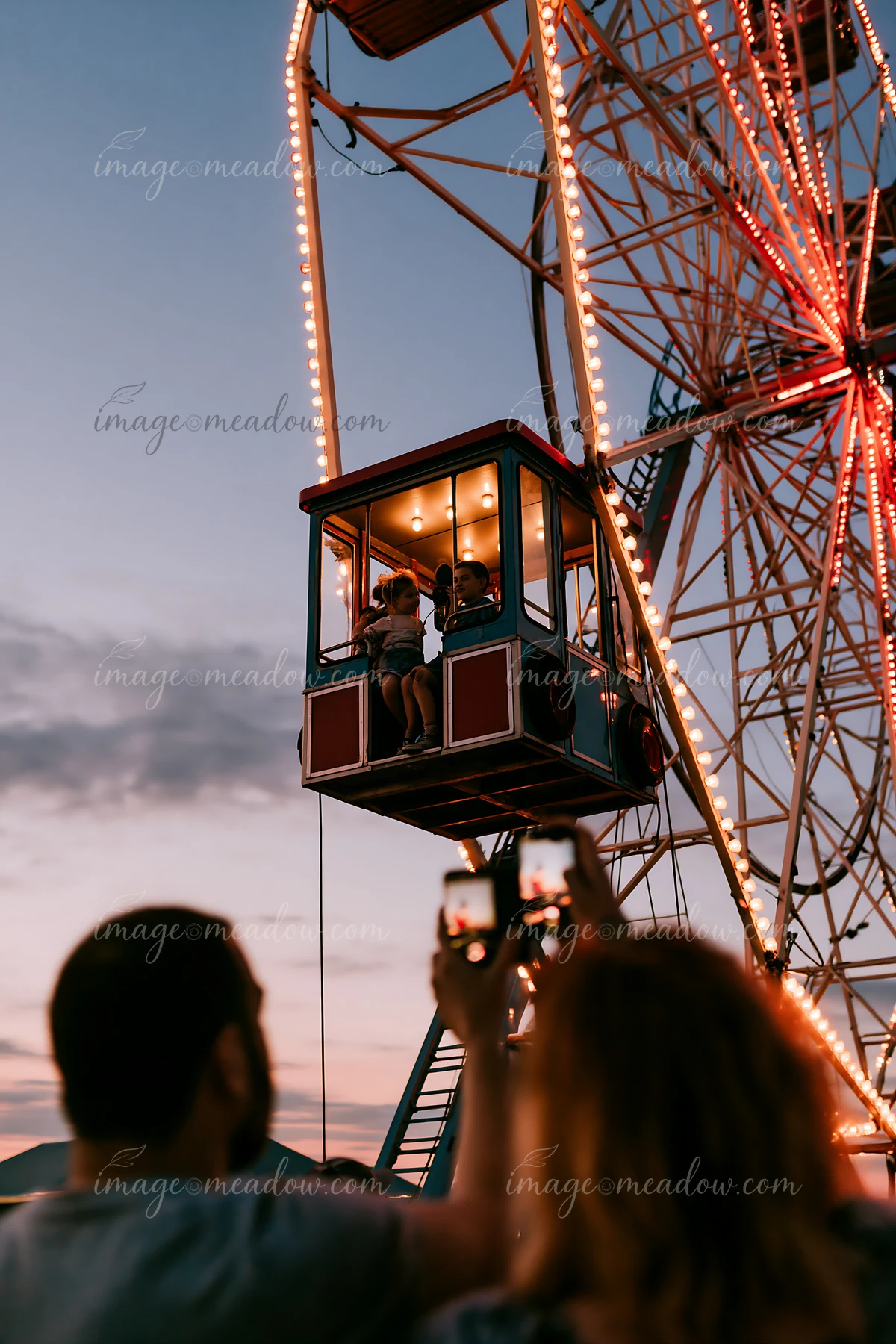 Parents photographing children on rides as they enjoy a ferris wheel at dusk.