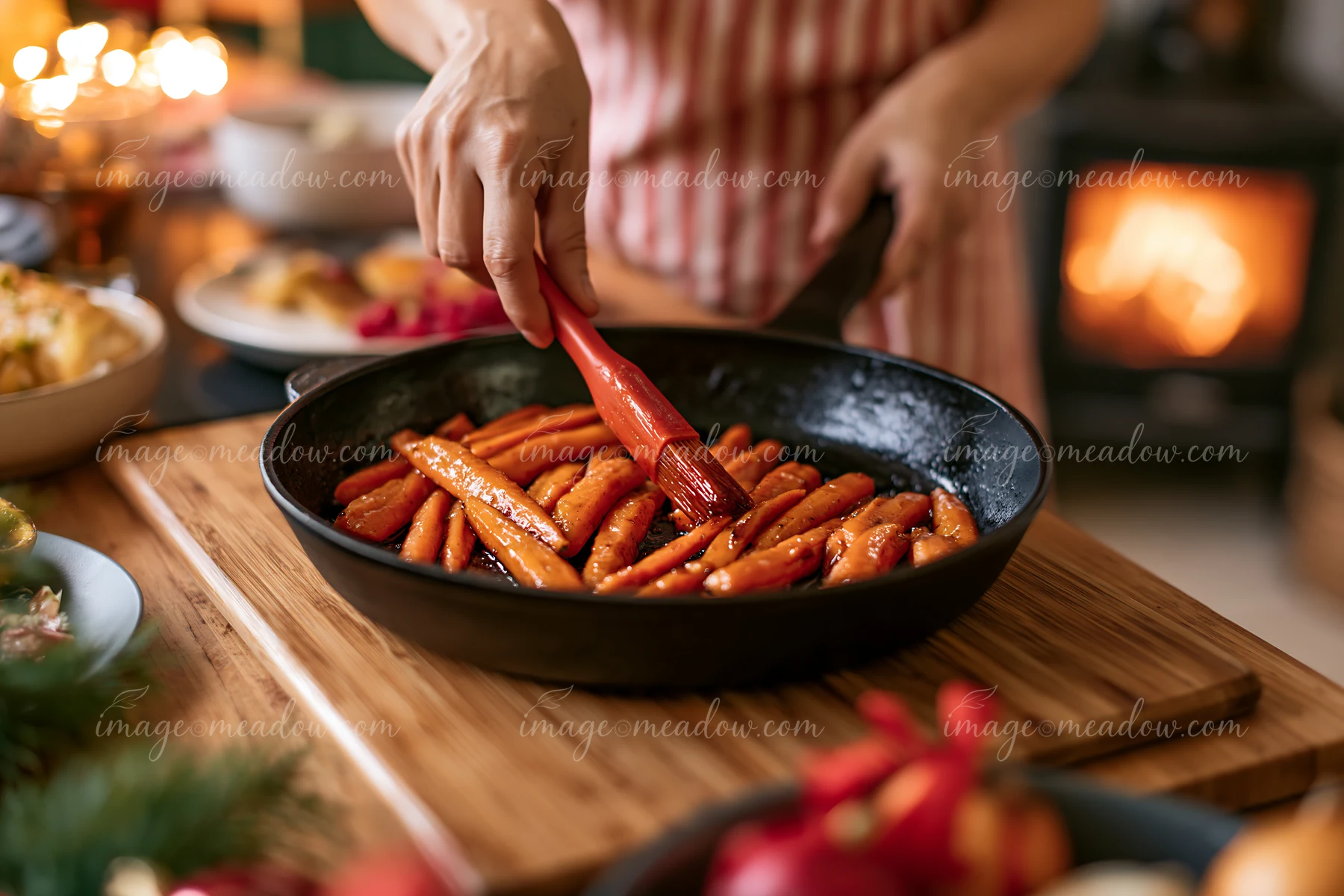Person brushes roasted carrots in a skillet on a wooden board in a warmly lit festive kitchen.