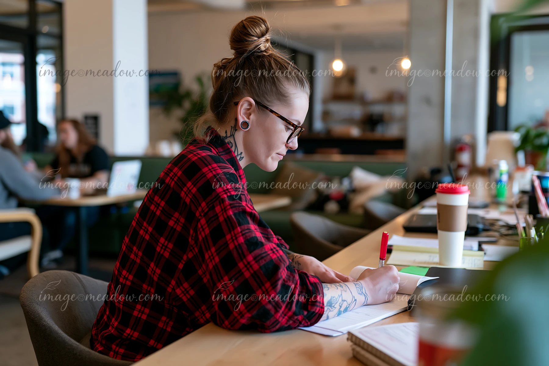 Person in a red plaid shirt writes in a notebook at a cafe desk beside a takeaway cup.