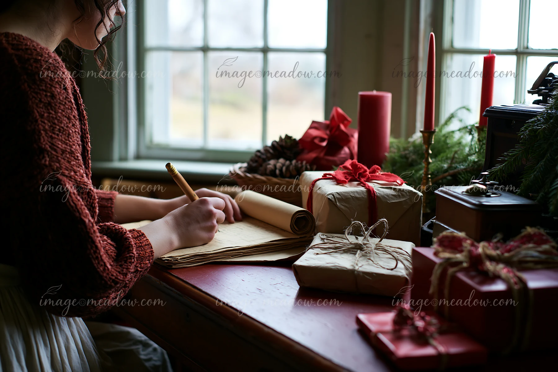 Person writes a gift list at a desk surrounded by wrapped presents, candles, and greenery.