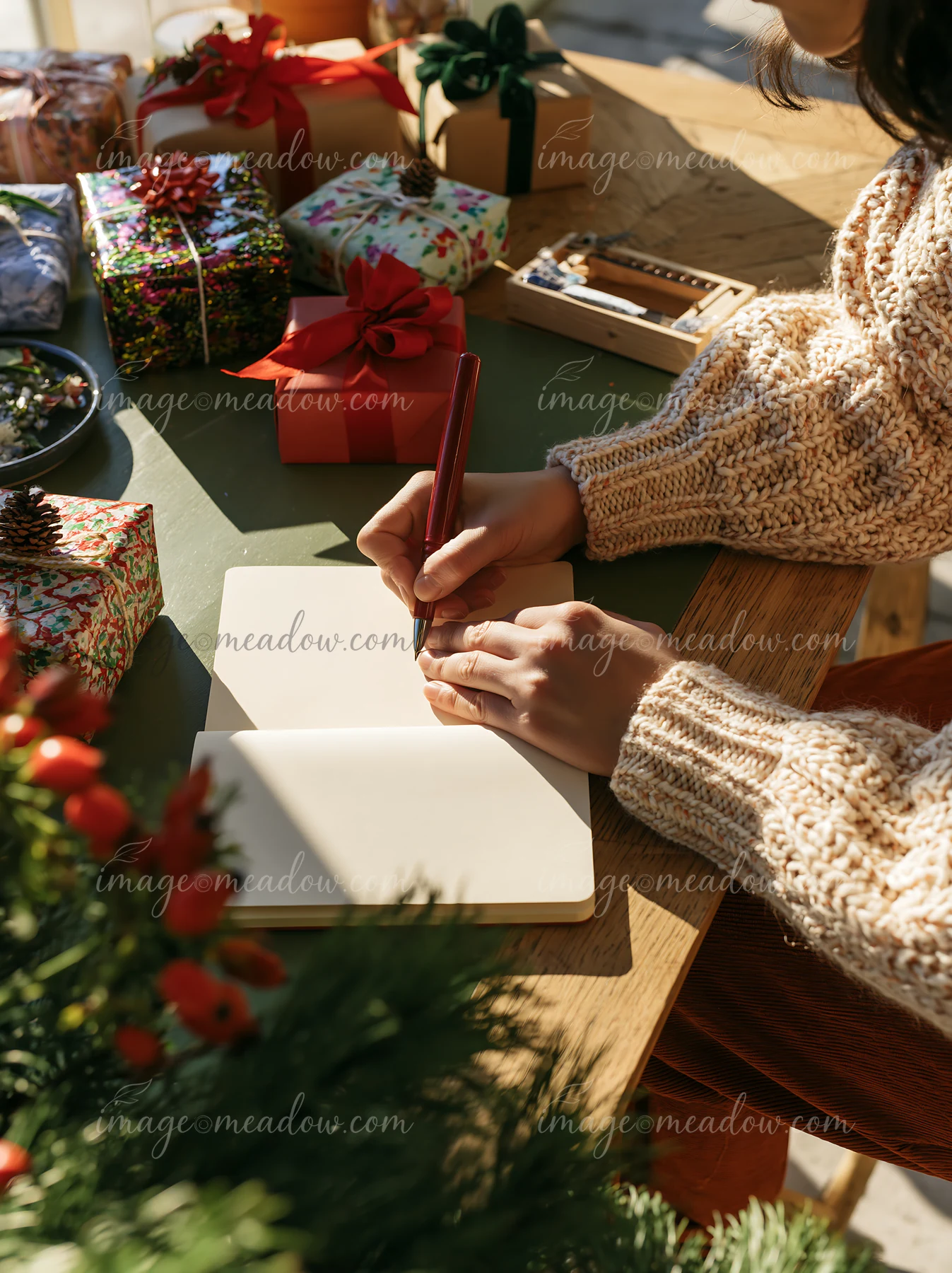 Person writing on a notebook at a desk surrounded by wrapped gifts in warm sunlight.