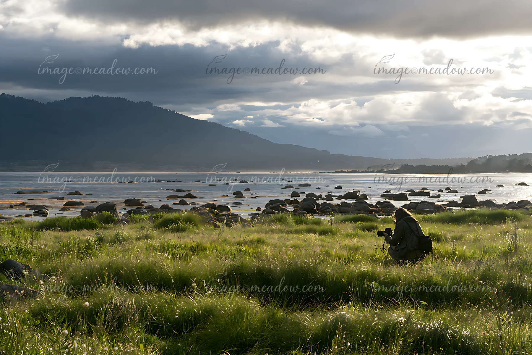 Photographer seated in a seaside meadow beside a rocky shore under dramatic clouds.
