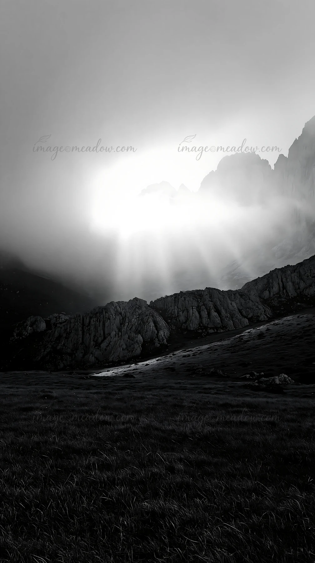 Rays of sunlight shine through clouds in the Picos de Europa mountain range. The scene captures grass and rocky terrain under a dramatic, cloudy sky during dayt