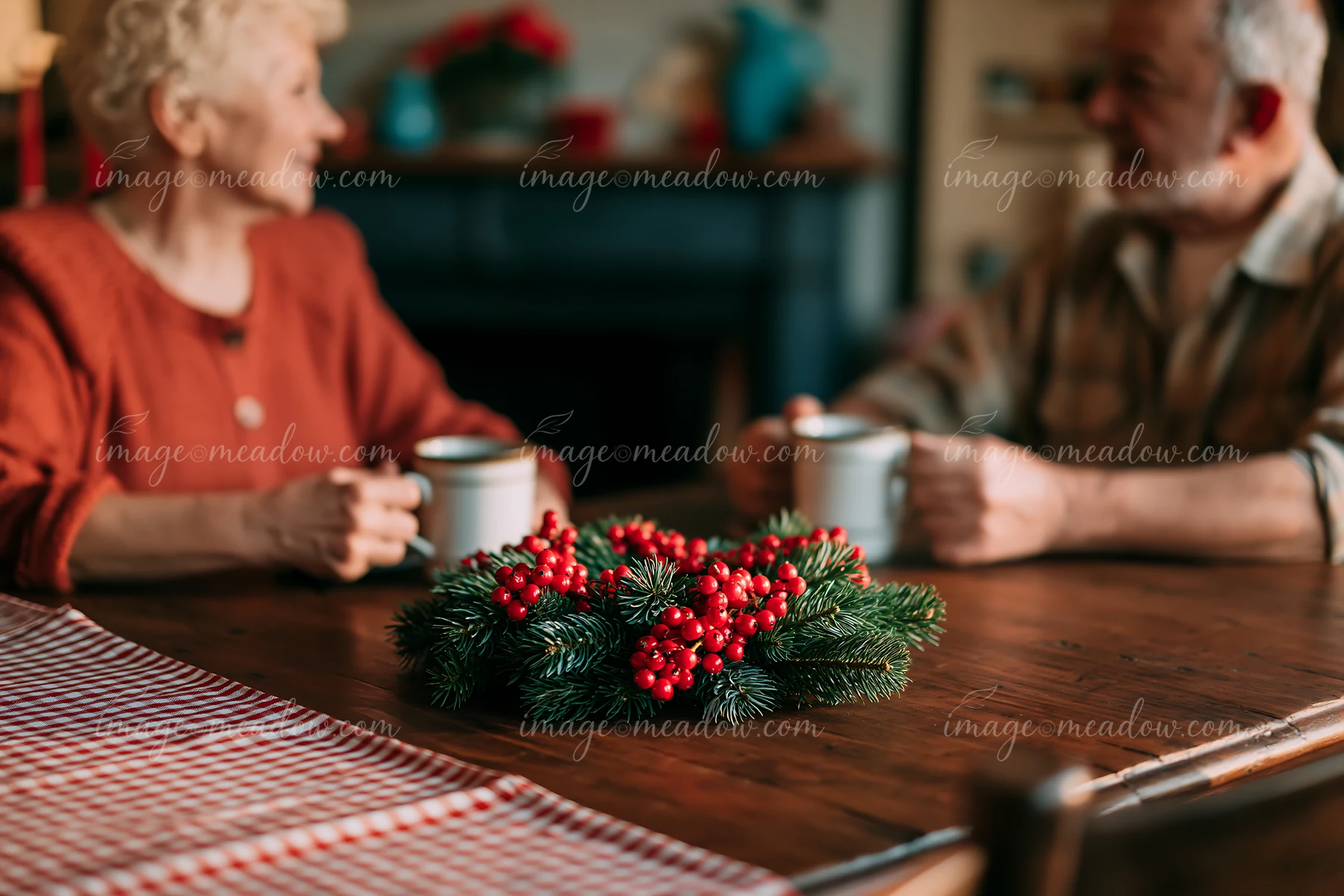 Pine wreath with red berries rests on a wooden dining table while an older couple holds mugs in the background.