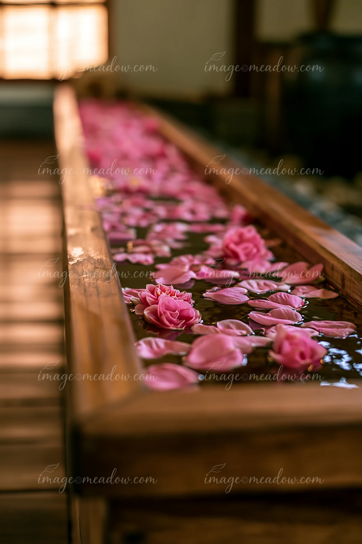 Pink flower petals and blossoms float in a narrow wooden water channel in warm, softly blurred surroundings.