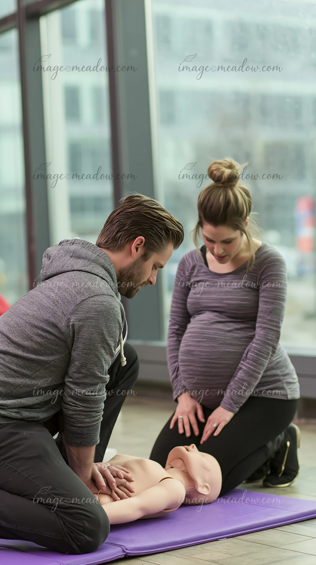 Pregnant couple practices chest compressions on an infant CPR training mannequin in a bright indoor class.