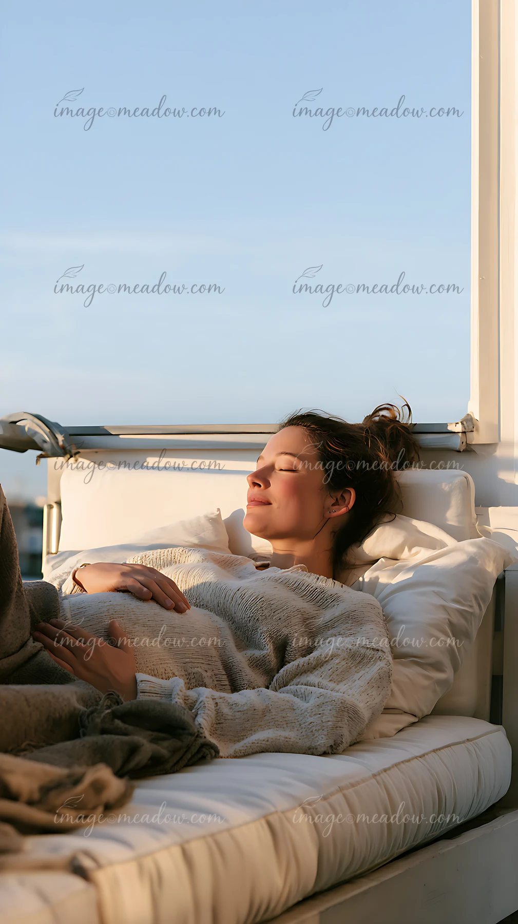 Pregnant woman rests with her hands on her belly in bed beside a sunlit window.