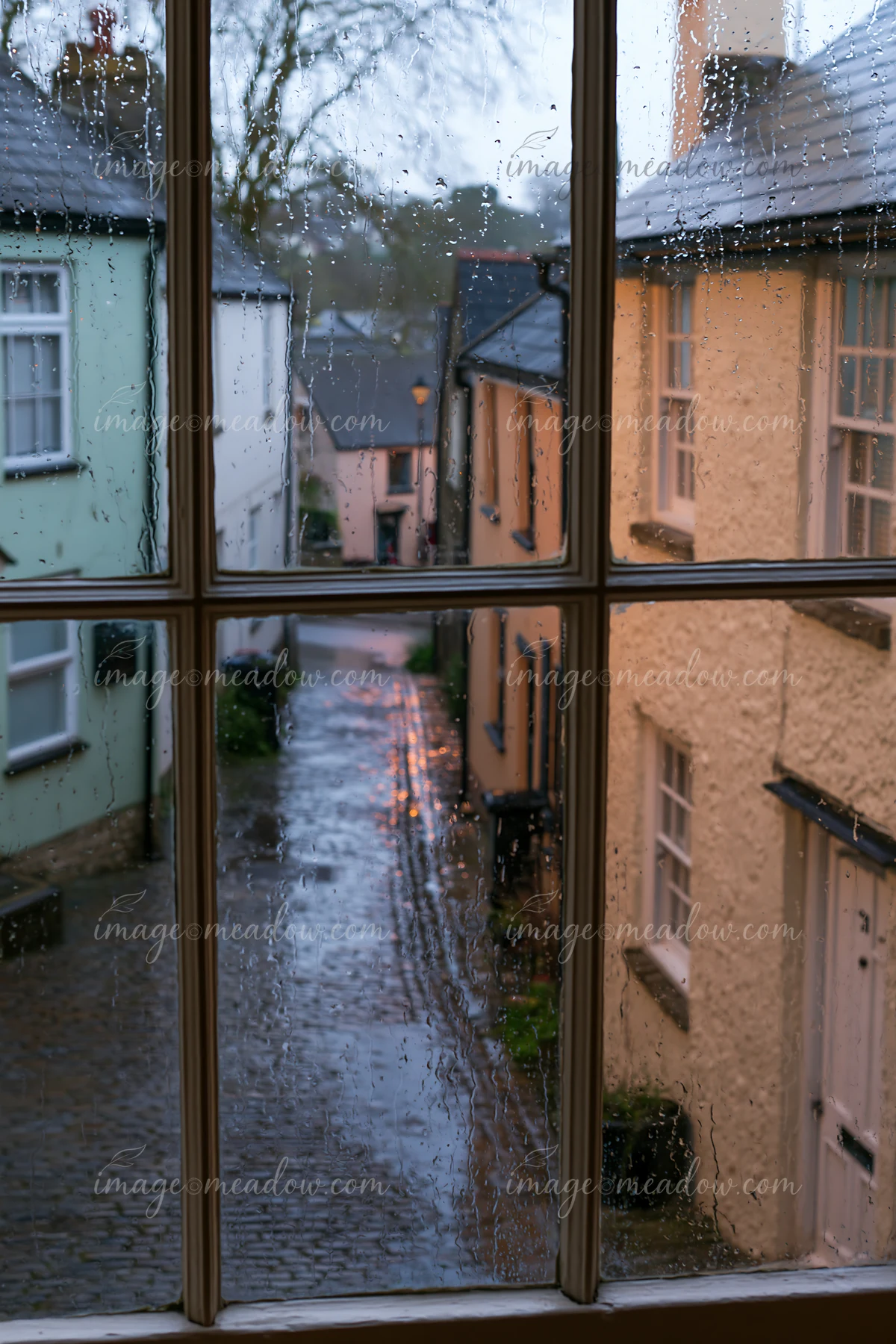 Raindrops are seen on a window, framing a view of a quaint cottage exterior.