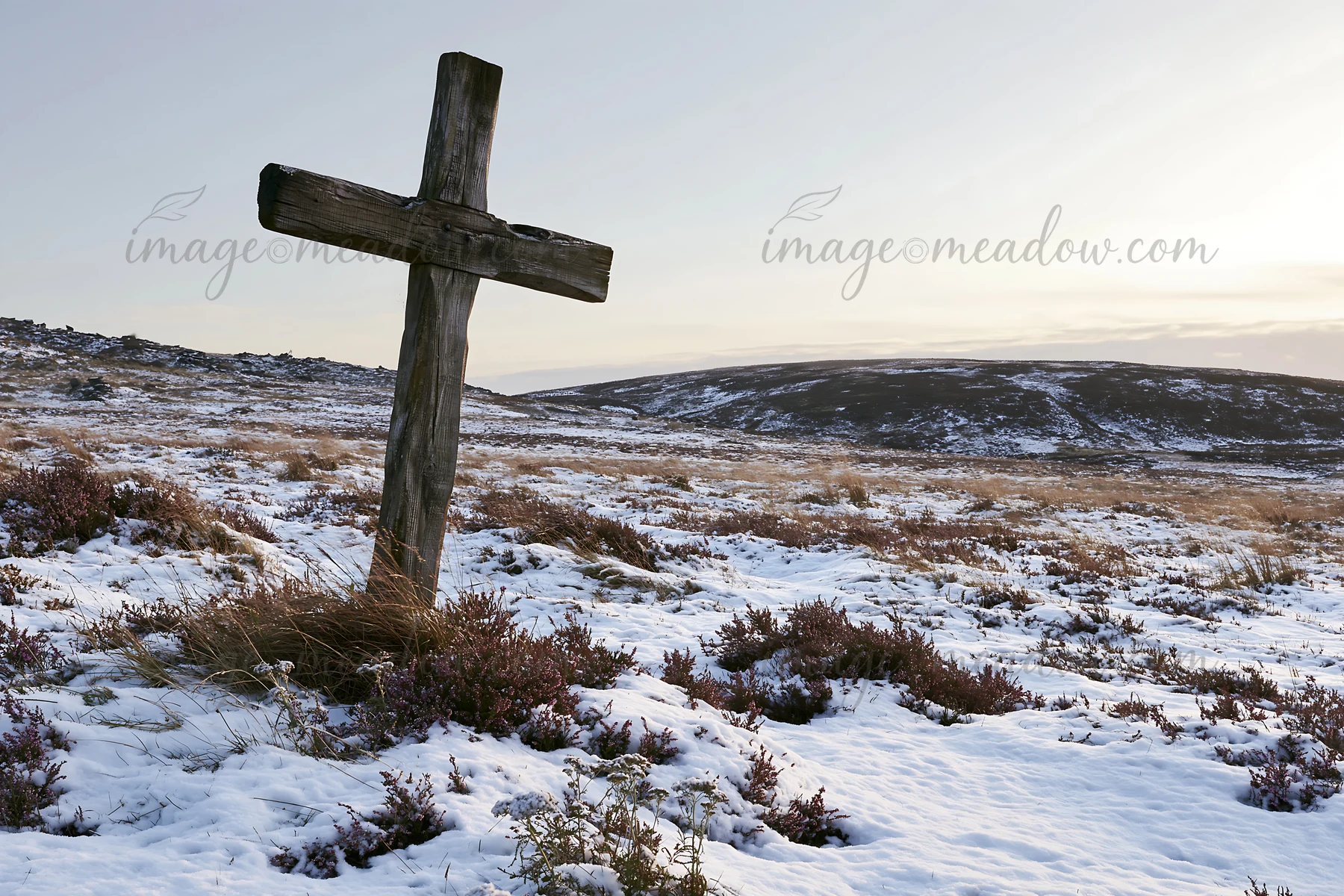 A wooden cross stands in a snow-covered meadow during winter. The landscape features dry grass and distant hills under a clear sky. This scene captures a quiet
