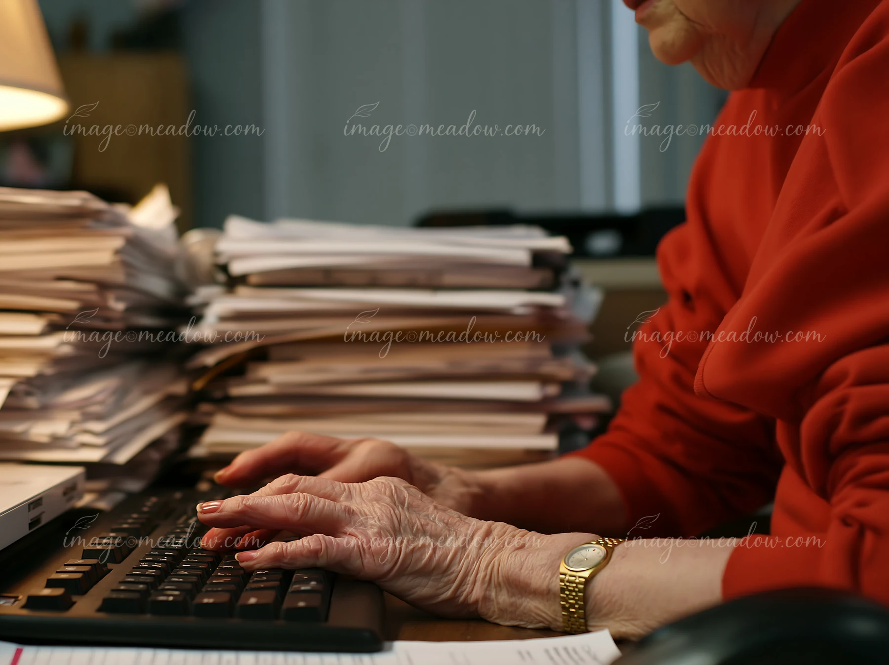 Senior woman typing with shaky hands at a cluttered desk surrounded by piles of paperwork.