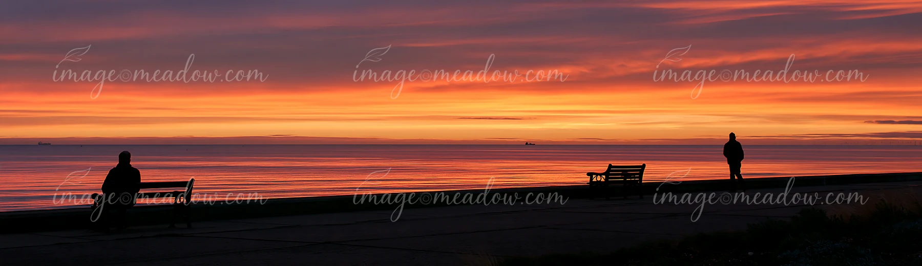 Silhouettes sit and stand beside seaside benches during a vivid orange sunset.