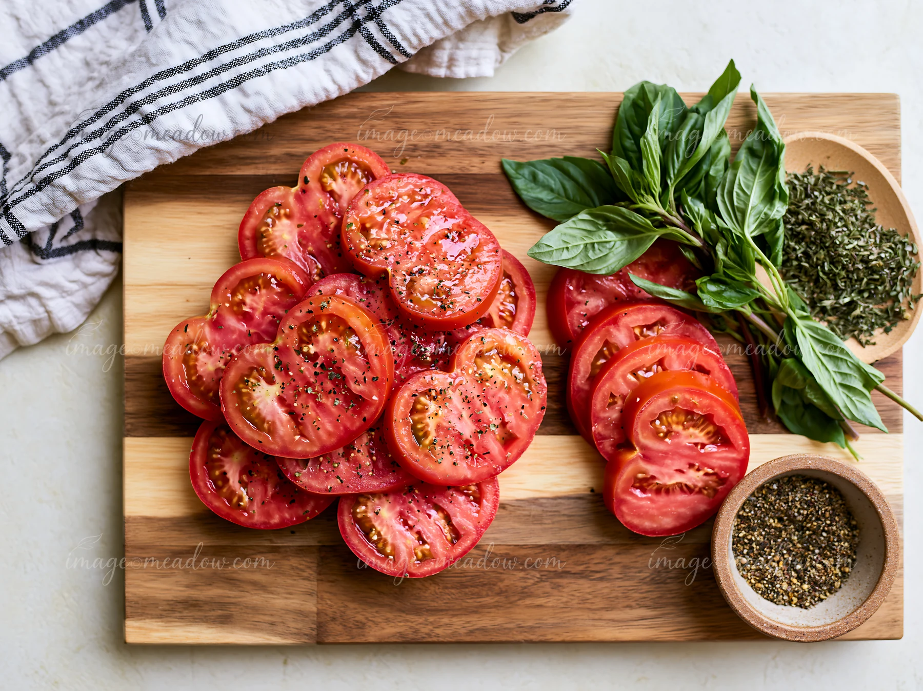 Sliced tomatoes, basil, and seasoning arranged on a wooden cutting board for cooking preparation.
