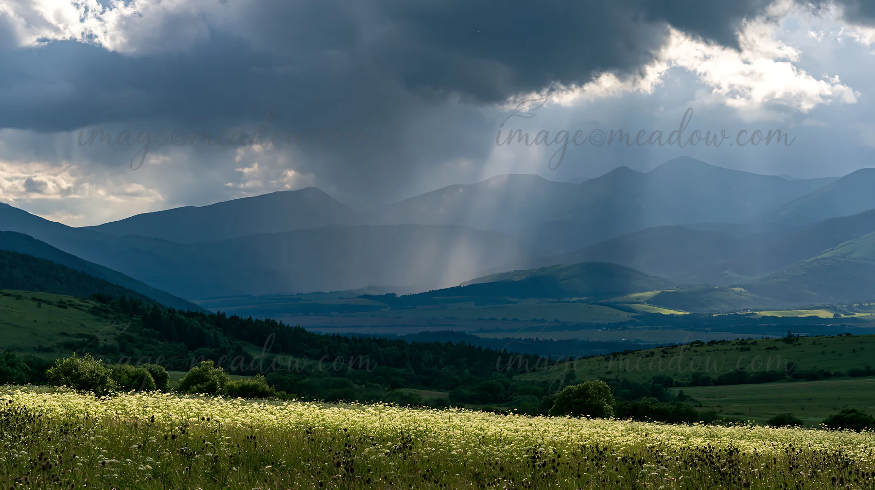 A view of a meadow in Slovakia across the Carpathian valley shows dark clouds and rays of sunlight. The landscape features green fields and distant mountains un