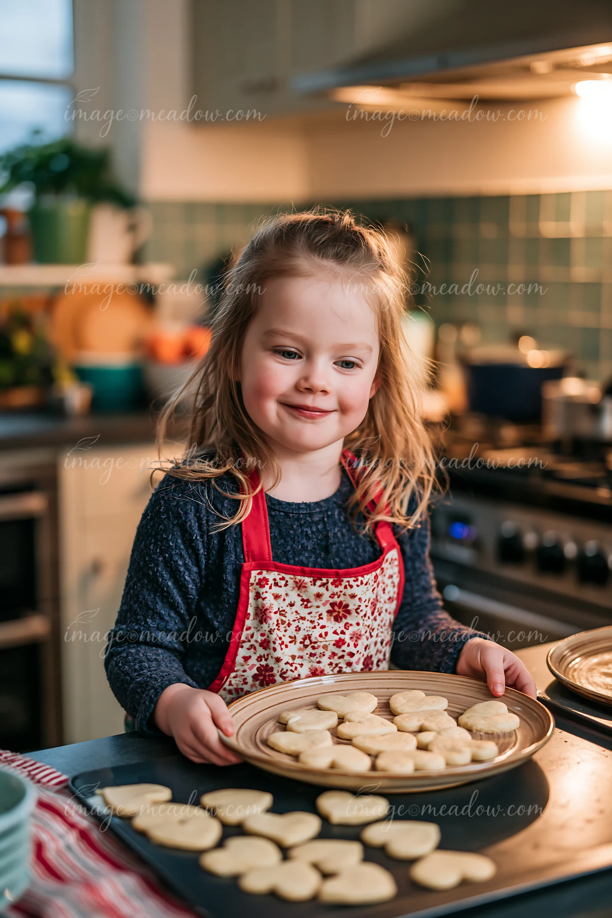Smiling child in a floral apron holds a tray of heart-shaped cookies in a warmly lit kitchen.